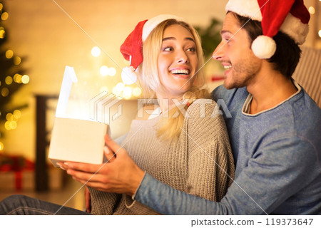 Excited couple opening magic xmas gift in living room, in front of Christmas tree Excited couple opening magic xmas gift in living room, in front of Christmas tree 119373647