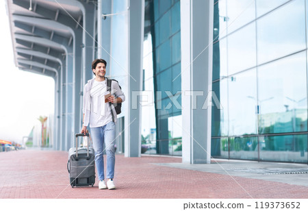 Going To Registration. Happy Young Man Walking With Suitcase And Backpack Near Airport Terminal, Holding Passport And Tickets In Hand, Free Space 119373652
