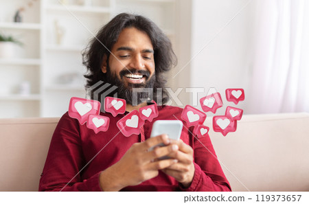 Indian man with long hair and a beard sits on a couch, smiling as he checks his smartphone. Around him are pink notification icons with white hearts, indicating that he has received many likes Indian man with long hair and a beard sits on a couch, smiling as he checks his smartphone. Around him are pink notification icons with white hearts, indicating that he has received many likes 119373657