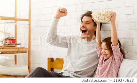 Cute emotional little girl and her father watching football match on TV, celebrating success of their team, sitting on bed, holding bowl with popcorn, family traditions concept Cute emotional little girl and her father watching football match on TV, celebrating success of their team, sitting on bed, holding bowl with popcorn, family traditions concept 119373658