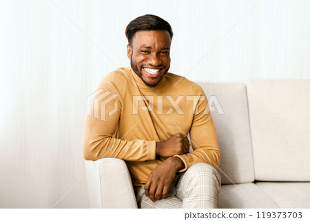 Millennial Afro Guy Smiling Looking At Camera Sitting On Sofa Indoor. Weekend At Home Millennial Afro Guy Smiling Looking At Camera Sitting On Sofa Indoor. Weekend At Home 119373703