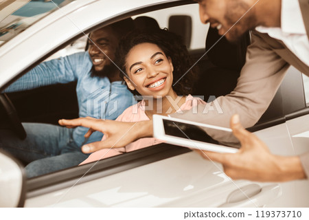 Happy Black Couple Buying New Car Testing And Talking With Salesman Sitting In Auto Dealership Showroom. Family Choosing Automobile. Happy Black Couple Buying New Car Testing And Talking With Salesman Sitting In Auto Dealership Showroom. Family Choosing Automobile. 119373710