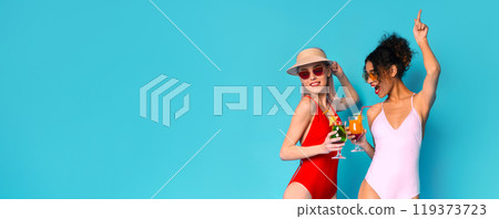 Two women in swimsuits are celebrating with drinks in their hands. They are standing in front of a bright blue background, panorama with copy space Two women in swimsuits are celebrating with drinks in their hands. They are standing in front of a bright blue background, panorama with copy space 119373723