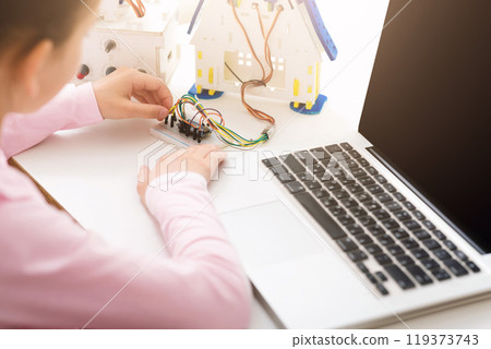 Girl is working on an electronic project at a desk, connecting wires to a breadboard. A small model house and a laptop are also on the desk, suggesting the project is related to robotics. Girl is working on an electronic project at a desk, connecting wires to a breadboard. A small model house and a laptop are also on the desk, suggesting the project is related to robotics. 119373743