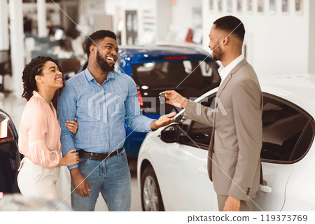 African American Couple Buying New Car Taking Key From Cheerful Salesman In Auto Dealership Showroom. Automobile Purchasing Service 119373769
