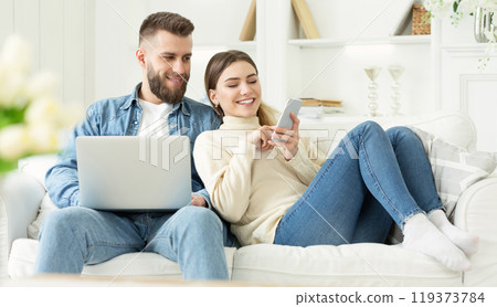 Millennial woman showing something on phone to husband, resting on sofa at home together 119373784