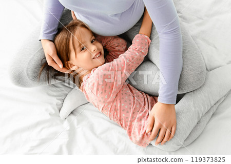 Mother love. Top view of pregnant young woman spending time with her little daughter, bonding on bed, girl hugging mommy tummy, copy space 119373825