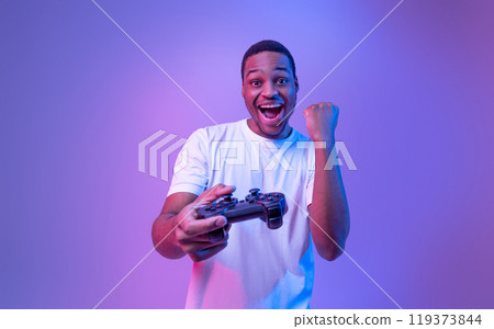 Video Gaming Concept. Euphoric African American Guy With Joystick Celebrating Game Win, Cheerful Black Man Raising Fist And Exclaiming With Excitement, Standing In Neon Light Over Purple Background Video Gaming Concept. Euphoric African American Guy With Joystick Celebrating Game Win, Cheerful Black Man Raising Fist And Exclaiming With Excitement, Standing In Neon Light Over Purple Background 119373844