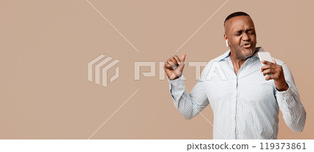 Modern entertainment. Emotional middle aged african american man in headphones singing at phone like a microphone, orange background Modern entertainment. Emotional middle aged african american man in headphones singing at phone like a microphone, orange background 119373861