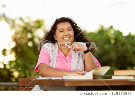 A young student sits at a wooden table surrounded by greenery, smiling while holding a pen and studying from an open book under the warm sunlight. A young student sits at a wooden table surrounded by greenery, smiling while holding a pen and studying from an open book under the warm sunlight. 119373875