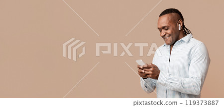Choosing next song. Cheerful african man in headphones using music mobile application, orange studio background Choosing next song. Cheerful african man in headphones using music mobile application, orange studio background 119373887
