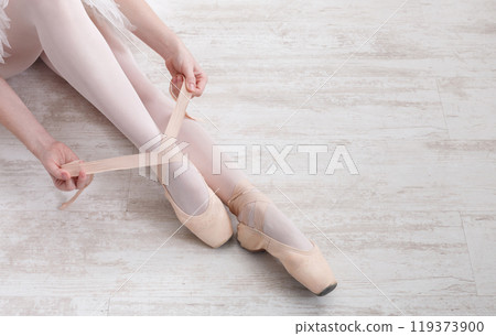 Beautiful legs of young ballerina who puts on pointe shoes at white wooden floor background, top view from above with copy space. Ballet practice. Beautiful slim graceful feet of ballet dancer. 119373900