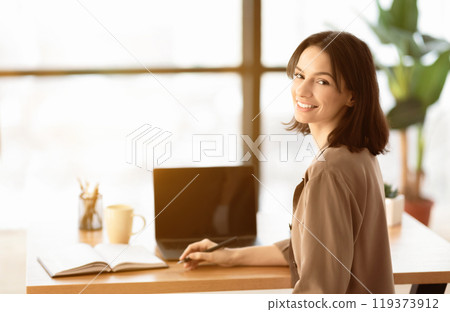 Happy Worker. Rear view of happy young woman looking over shoulder and smiling, sitting at her workplace with laptop Happy Worker. Rear view of happy young woman looking over shoulder and smiling, sitting at her workplace with laptop 119373912
