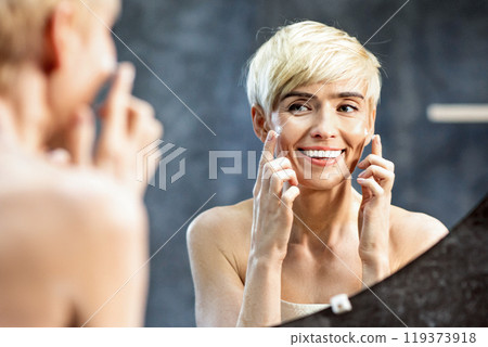 Mature Lady Moisturizing Skin Applying Facial Cream Looking In Mirror Standing In Bathroom At Home. Selective Focus Mature Lady Moisturizing Skin Applying Facial Cream Looking In Mirror Standing In Bathroom At Home. Selective Focus 119373918