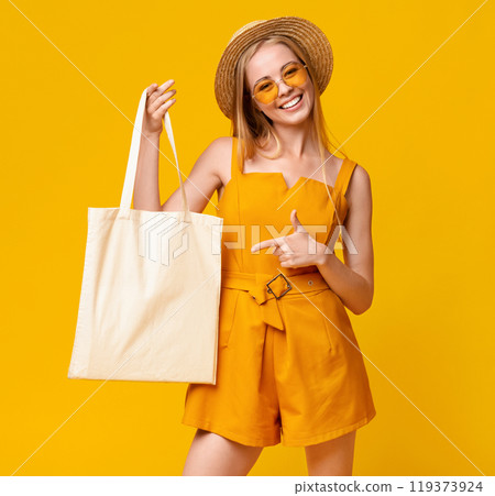 Mockup For Design. Cheerful Summer Girl Holding White Eco Tote Bag And Pointing At It, Posing Over Yellow Background With Empty Space Mockup For Design. Cheerful Summer Girl Holding White Eco Tote Bag And Pointing At It, Posing Over Yellow Background With Empty Space 119373924