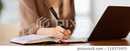 Taking Notes. Young secretary sitting at desk with laptop computer and talking on mobile phone, writing in notebook 119373935