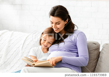 Friendly intelligent family pregnant mother and little daughter reading book on couch at home, empty space 119373969