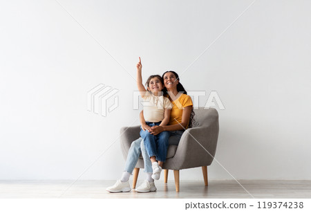 Check This. Happy Young Mother And Little Daughter Sitting In Armchair And Pointing Up, Cheerful Middle Eastern Mom And Female Child Demonstrating Copy Space Above Their Heads While Resting In Chair 119374238
