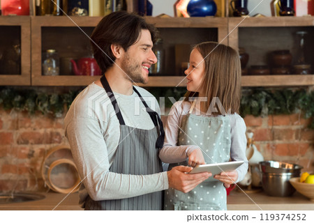 Smiling little girl and her young dad using digital tablet at kitchen, checking cookies recipe, enjoying spending time together 119374252