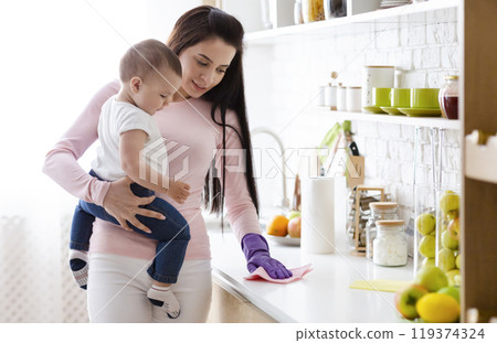 Domestic chores at maternity leave. Young mother with baby boy cleaning kitchen at home, free space Domestic chores at maternity leave. Young mother with baby boy cleaning kitchen at home, free space 119374324