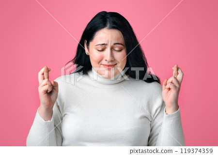 Cherished wish. Portrait of young overweight woman pleading for something with crossed fingers and closed eyes over pink background 119374359