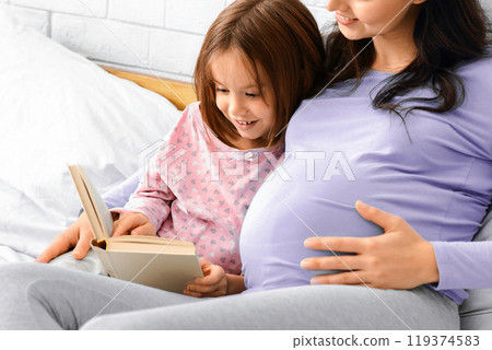 Pregnant lady reading fairy tales to her little cute daughter, spending time together at home, lying in bed, copy space Pregnant lady reading fairy tales to her little cute daughter, spending time together at home, lying in bed, copy space 119374583