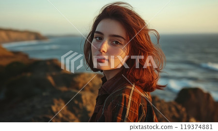 A young woman stands on a rocky cliff, looking out over the ocean's waves during golden hour. Her hair is gently tousled by the breeze as she enjoys the serene coastal landscape. 119374881
