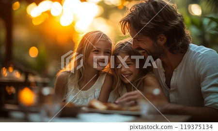 A father smiles warmly as he enjoys a light-hearted dinner with his two daughters, surrounded by a glowing sunset and lush greenery. 119375134