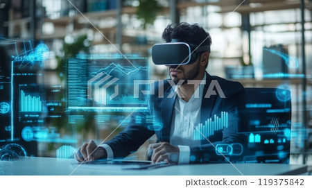 A man wearing a virtual reality headset sits at a desk in an office. The headset displays various graphs and data visualizations 119375842