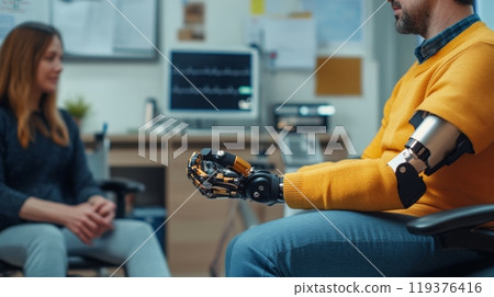 In a warm clinic, a man wearing a yellow sweater showcases his advanced prosthetic arm while speaking with a therapist seated nearby In a warm clinic, a man wearing a yellow sweater showcases his advanced prosthetic arm while speaking with a therapist seated nearby 119376416