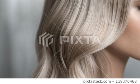Silky, wavy blonde locks cascade gently over the shoulder, illuminated by soft, natural light, highlighting the hair's shine and texture. 119376468