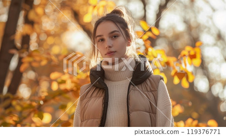 A young woman stands gracefully in a park wearing a cozy sweater and a puffy vest, surrounded by vibrant autumn foliage. 119376771