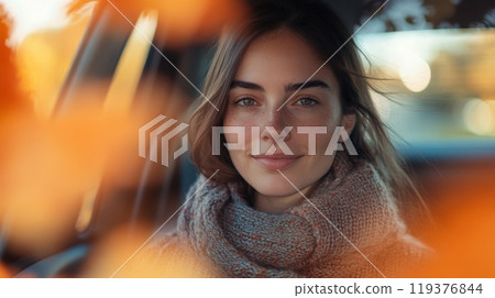 A young woman is enjoying a moment inside her car, wearing a cozy scarf and smiling gently. Colorful autumn leaves frame her as the warm sunset light adds a soft glow to the scene. A young woman is enjoying a moment inside her car, wearing a cozy scarf and smiling gently. Colorful autumn leaves frame her as the warm sunset light adds a soft glow to the scene. 119376844