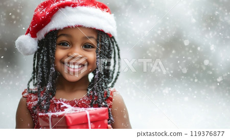 A smiling child wearing a festive Santa hat stands in the snow, holding a beautifully wrapped Christmas gift. The joy of the season is evident as snowflakes gently fall around her. 119376877