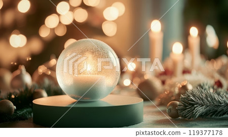A snow globe containing a burning candle rests on a circular base. The snow globe sits on a table with other Christmas decorations, including an artificial evergreen sprig and a golden bauble. 119377178