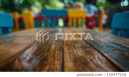 A close-up view of a weathered wooden table with blurred colorful chairs in the background. 119377519