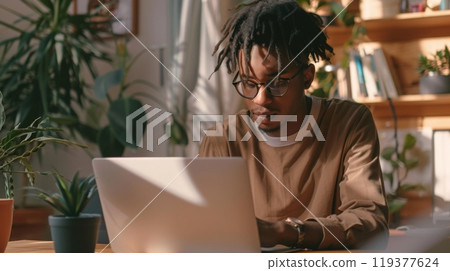 A young man with dreadlocks concentrates on his laptop in a bright home office filled with plants. A young man with dreadlocks concentrates on his laptop in a bright home office filled with plants. 119377624