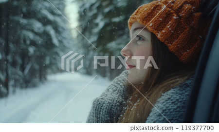 A woman with a warm hat gazes thoughtfully out of the car window on a snowy road surrounded by evergreen trees. The serene atmosphere of the winter landscape creates a sense of tranquility. 119377700
