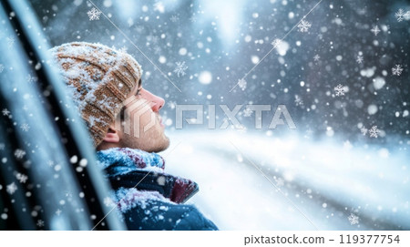 A young man relaxes against a vehicle, gazing upwards as delicate snowflakes drift down around him. The tranquil winter scene captures the beauty of falling snow in a serene environment. 119377754