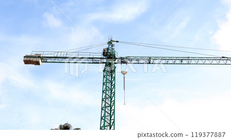Construction crane tower rises above the skyline with workers maneuvering equipment at a busy urban site during daylight hours 119377887
