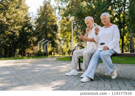 Low-angle view of pretty senior couple with white hair happily relaxing in city park, sitting on bench,enjoying smartphones on beautiful sunny day. Concept of happy retirement life. Low-angle view of pretty senior couple with white hair happily relaxing in city park, sitting on bench,enjoying smartphones on beautiful sunny day. Concept of happy retirement life. 119377938