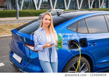 A woman stands by her electric car, charging while holding a green bottle and using her phone to pay. Green energy concept A woman stands by her electric car, charging while holding a green bottle and using her phone to pay. Green energy concept 119384039