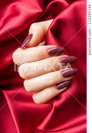 Elegant hand with burgundy nails resting on soft red satin fabric in a luxurious indoor setting 119385144