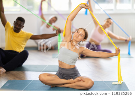 Girl conducts Pilates multinational group sitting with elastic band in hands above head body bends Girl conducts Pilates multinational group sitting with elastic band in hands above head body bends 119385296