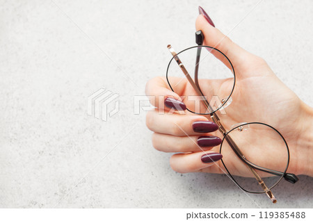 A close-up of a hand holding stylish glasses with burgundy nails on a light textured surface 119385488