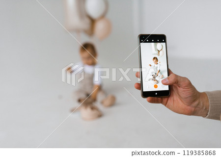 Father takes pictures of his son on his smartphone. Baby boy is sitting on the floor and eating a birthday cake 119385868