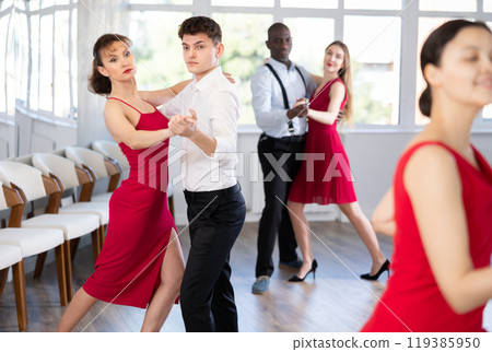 Pretty young woman dancing waltz with partner at group of multinational people in ballroom dancing class 119385950