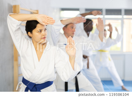 Middle-aged woman attendee of karate classes practicing kata standing in row with others Middle-aged woman attendee of karate classes practicing kata standing in row with others 119386104