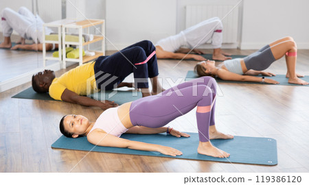 Asian woman performs glute bridge during Pilates multinational training with elastic band on shins Asian woman performs glute bridge during Pilates multinational training with elastic band on shins 119386120
