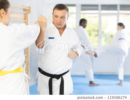 Middle-aged man attendee of karate classes fighting with his opponent in sports hall Middle-aged man attendee of karate classes fighting with his opponent in sports hall 119386274